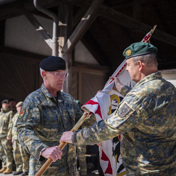 Zwei Soldaten in Uniform übergeben eine Fahne vor einer Gruppe von Personen.