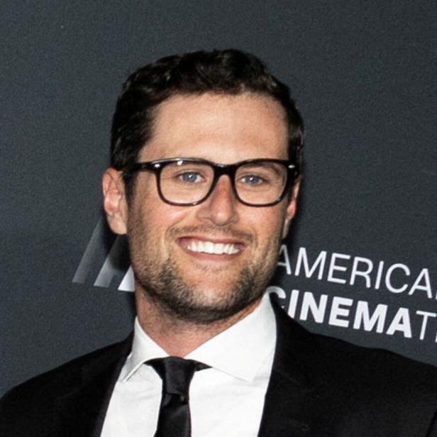 Ein Mann mit Brille, Anzug und Krawatte lächelt vor einer dunklen Wand mit weißer Schrift.