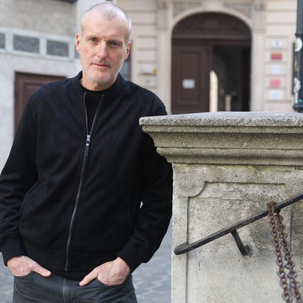 Ein Mann mit grauem Haar und Bart steht in schwarzer Jacke an einer steinernen Brüstung in einer Altstadtgasse.