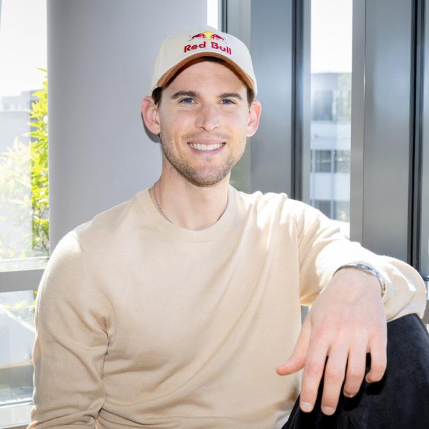 Ein junger Mann mit beigem Pullover und Red Bull-Kappe sitzt lächelnd vor einem Fenster mit modernem Rahmen.