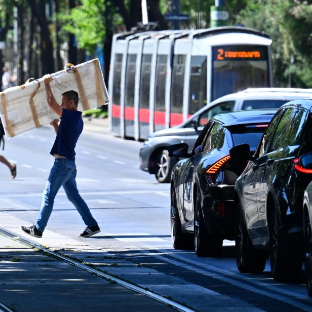 Ein Mann geht über die Straße, Autos warten, im Hintergrund fährt eine Straßenbahn.