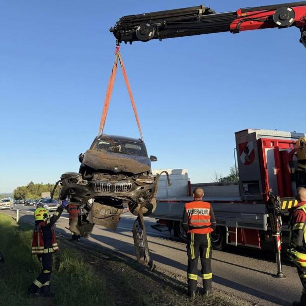 Ein stark beschädigtes Auto wird von einem Kran angehoben, während Feuerwehrleute daneben stehen.