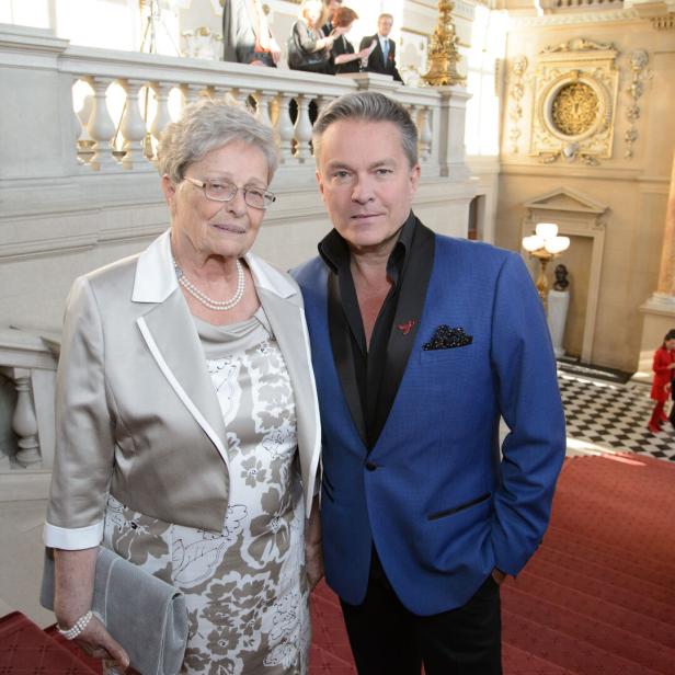 Eine ältere Frau und ein Mann im Anzug stehen auf einer prunkvollen Treppe in einem festlichen Gebäude.