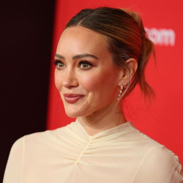 Hilary Duff mit hochgesteckten Haaren und Ohrringen vor rotem Hintergrund.