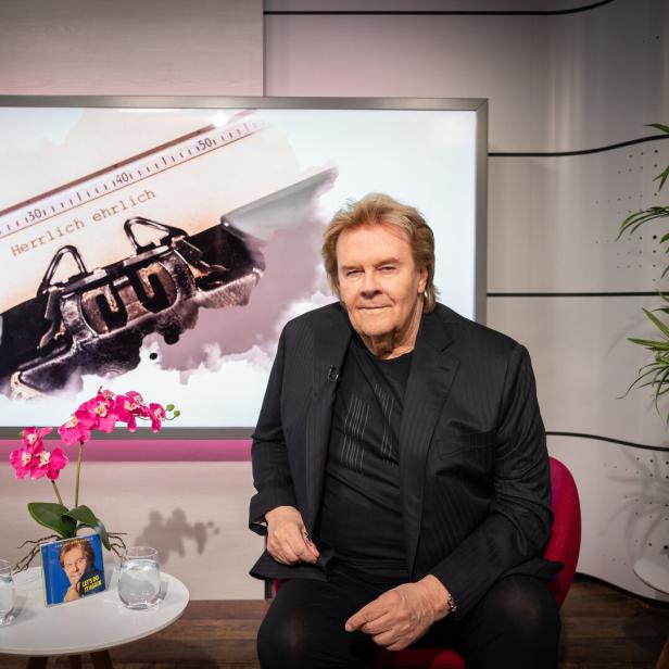 Howard Carpendale mit blondem Haar und schwarzem Sakko lächelt vor einem hellen Hintergrund.