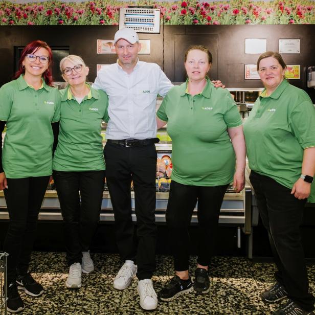 Fünf Personen in Arbeitskleidung stehen lächelnd vor einer Fleischtheke in einem Supermarkt nebeneinander.