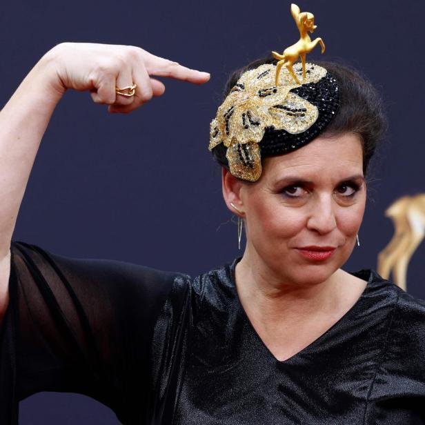 Frau mit schwarzem Kleid zeigt auf einen auffälligen Hut, der mit goldener Hirschfigur und Glitzer verziert ist.