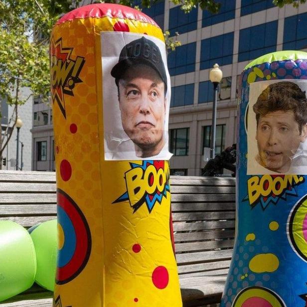 Auf einer Parkbank stehen zwei bunte Boxsäcke mit den Gesichtern von Elon Musk und einem weiteren Mann.