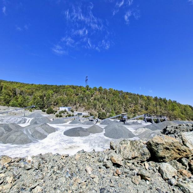 Steinbruch mit mehreren Kieshügeln, Förderbändern und angrenzendem Wald unter klarem, blauem Himmel.