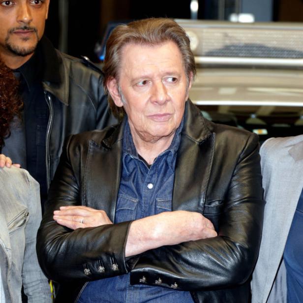 Jan Fedder mit verschränkten Armen in Jeans und Lederjacke, umgeben von anderen Personen, vermutlich bei einer Veranstaltung.