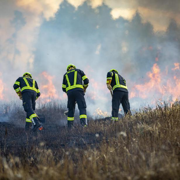 Drei Feuerwehrleute bekämpfen einen Flächenbrand auf einem trockenen Feld, während dichter Rauch und Flammen aufsteigen.
