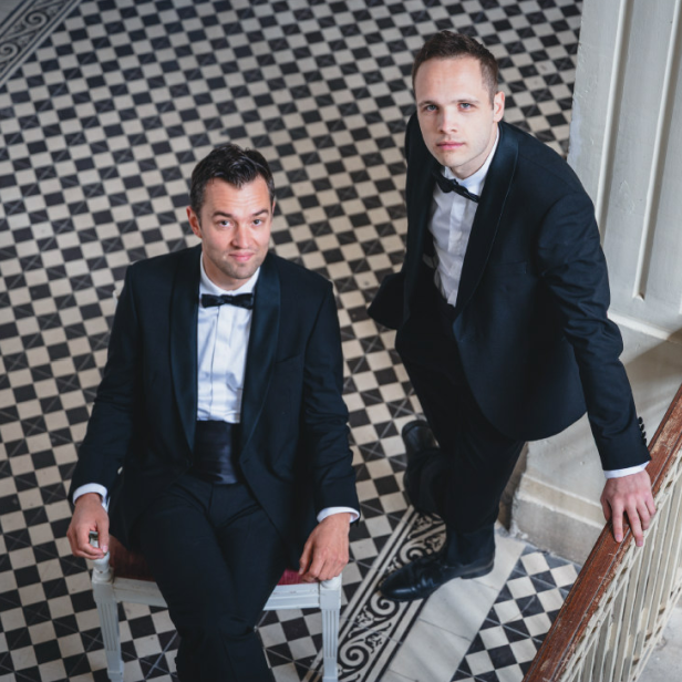 Nils Strunk und Lukas Schrenk im eleganten Anzug auf einer historischen Treppe, verantwortlich für die Neuinterpretation von „Die Fledermaus“ bei den Festspielen Reichenau 2026.