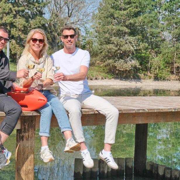 Drei Personen sitzen lachend auf einem Steg am Wasser, stoßen mit Gläsern an und tragen Sonnenbrillen.