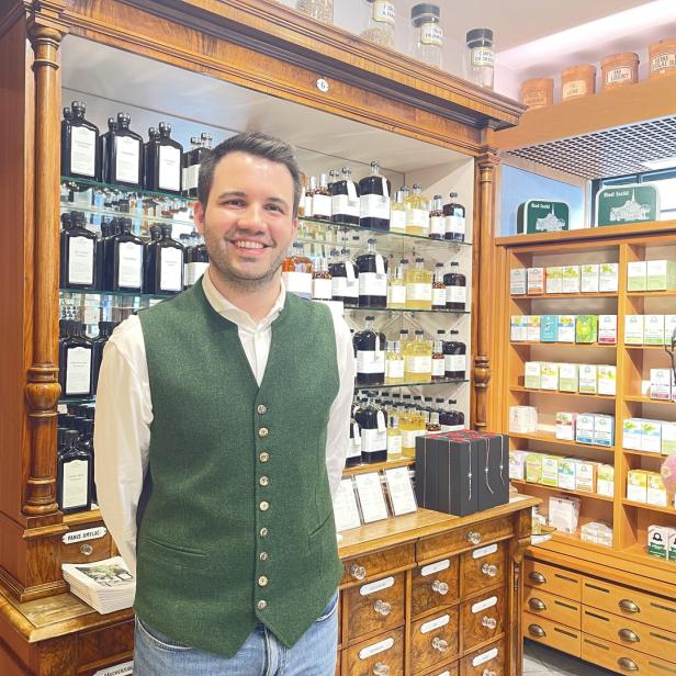 Ein Mann mit grünem Gilet steht lächelnd in einer traditionellen Apotheke vor Regalen mit Flaschen und Produkten.