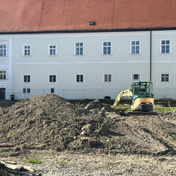 Ein Bagger steht auf einer Baustelle vor einem großen, weißen Gebäude mit rotem Dach.