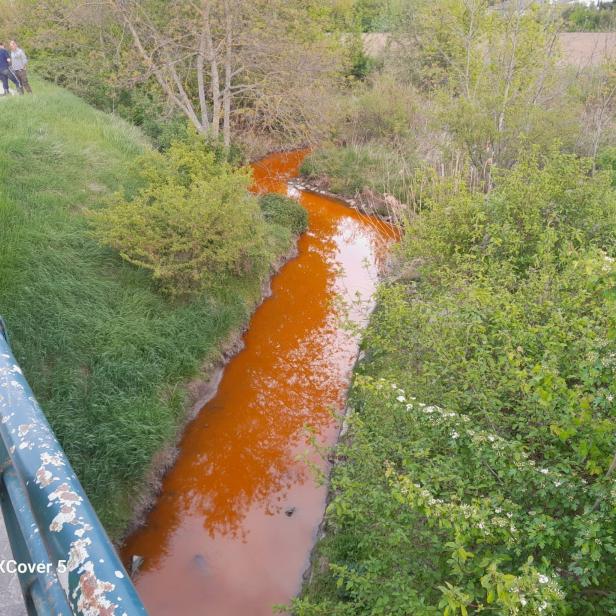 Oranger Fluss schlängelt sich durch grüne Vegetation, mehrere Personen stehen am Ufer.