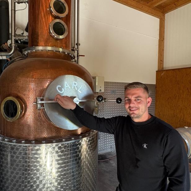 Ein Mann steht neben einer kupfernen Destille und hält den Griff einer runden Tür mit der Aufschrift „CARL“.