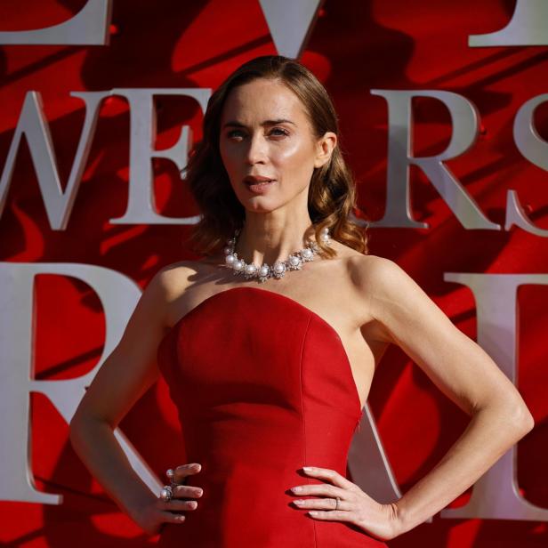 Emily Blunt mit Perlenkette und rotem, trägerlosem Kleid posiert vor rotem Hintergrund.