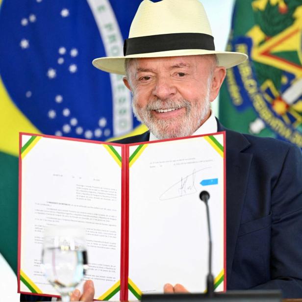 Ein älterer Mann mit Hut hält ein unterzeichnetes Dokument vor sich, im Hintergrund sind brasilianische Flaggen zu sehen.
