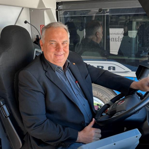 Ein lächelnder Mann im Anzug sitzt am Steuer eines Busses.