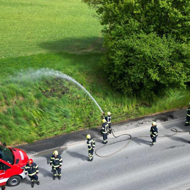 Mehrere Feuerwehrleute löschen mit einem Schlauch einen kleinen Brand am Straßenrand neben zwei Einsatzfahrzeugen.