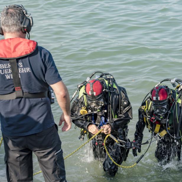 Zwei Feuerwehrtaucher steigen mit Ausrüstung aus dem Wasser, eine Person mit Headset steht am Ufer.