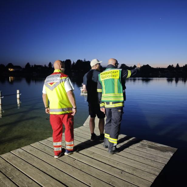 Drei Personen stehen auf einem Steg am Wasser, zwei davon in Einsatzkleidung, während die Dämmerung einsetzt.