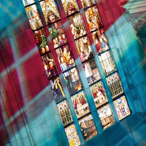 Buntes Licht fällt durch ein großes Kirchenfenster mit detailreichen Glasmalereien und Figuren auf steinerne Wände.