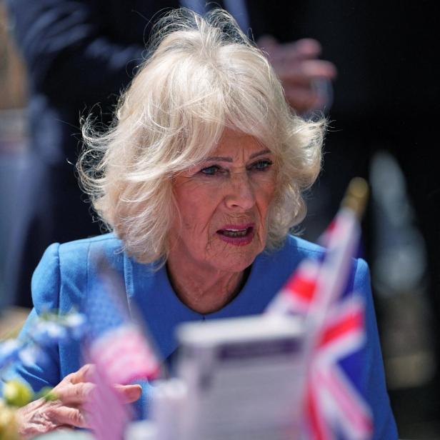 Königin Camilla, in einem blauen Blazer, spricht vor britischen Flaggen.