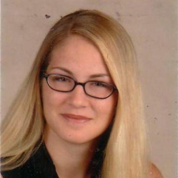 Junge Frau mit langen blonden Haaren und Brille, trägt ein dunkles Oberteil und lächelt leicht vor neutralem Hintergrund.
