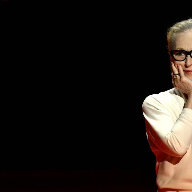 Meryl Streep mit Brille und hellem Oberteil posiert vor dunklem Hintergrund.