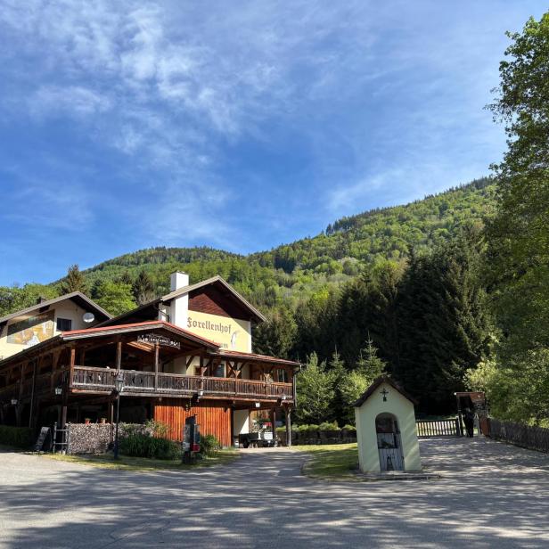 Ein Gasthaus mit Holzveranda steht vor bewaldeten Hügeln, daneben eine kleine Kapelle und umgeben von grünen Bäumen.