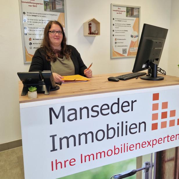 Eine Frau steht hinter einem Schild mit "Manseder Immobilien" 