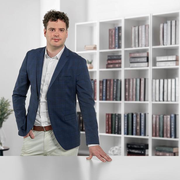 Mann im Anzug steht in modernem Büro mit Bücherregal und Pflanzen im Hintergrund.