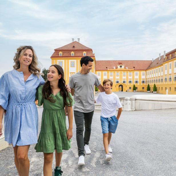 Vier Personen spazieren lachend vor dem gelben Schloss Hof und einer steinernen Löwenskulptur.