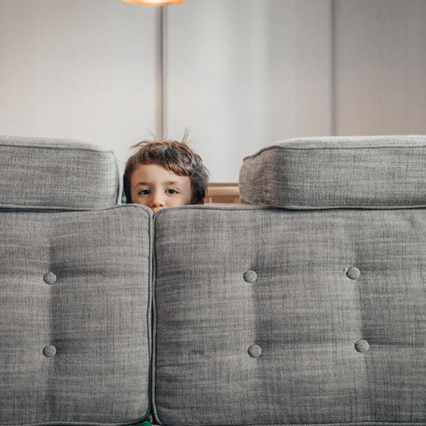 Ein Kind blickt neugierig über die Rückenlehne eines grauen Sofas, das im Mittelpunkt eines modern eingerichteten Wohnzimmers steht.