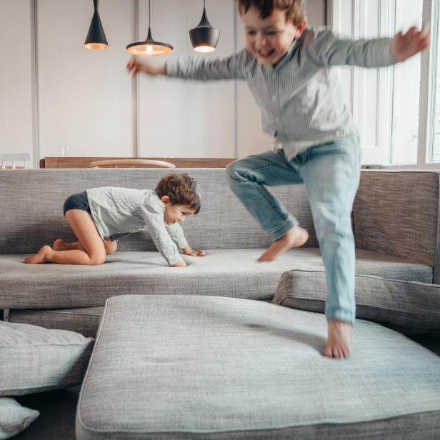 Zwei Kinder toben auf einem grauen Sofa mit abgenommenen Polstern in einem hellen Wohnzimmer, was die Alltagstauglichkeit und Robustheit des Sofas verdeutlicht.