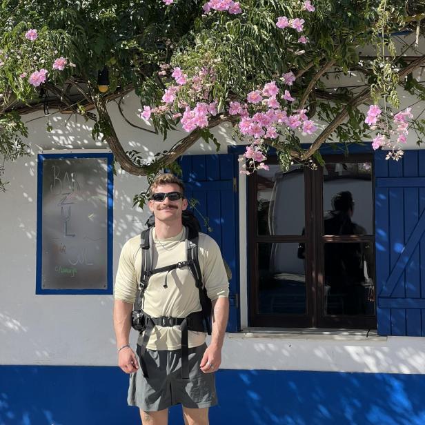 Ein junger Mann steht unter einem blühenden Baum, mit Rucksack, kurzer Hose und Sonnenbrille.