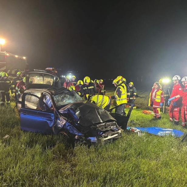 Mehrere Einsatzkräfte stehen nachts um ein stark beschädigtes Auto auf einer Wiese, Rettungsfahrzeuge und Scheinwerfer im Hintergrund.