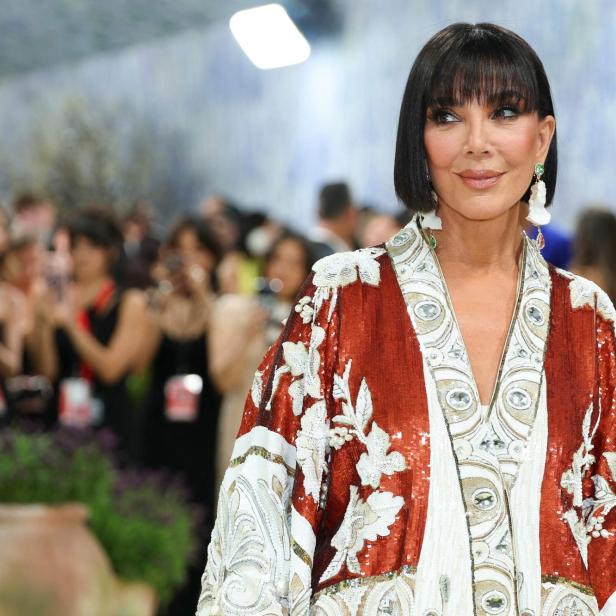 Kris Jenner trägt eine verzierte, rot-weiße Robe und blickt in einer Menschenmenge zur Seite.