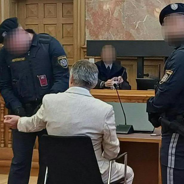 Ein Mann in einem hellen Anzug sitzt im Gerichtssaal, bewacht von zwei Justizwachebeamten.