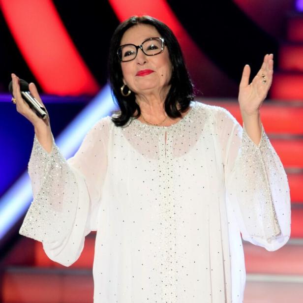 Nana Mouskouri mit Brille und weißer Bluse hält ein Mikrofon in der Hand.