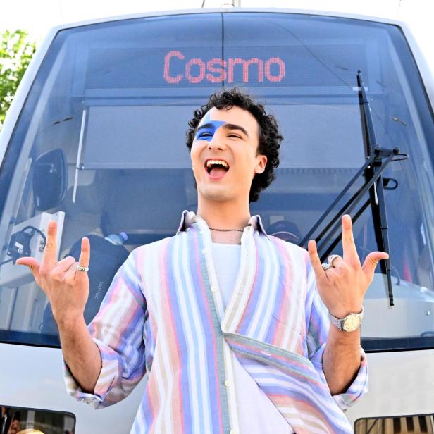 Eine Person mit blauem Gesichtsmakeup und bunter Kleidung steht lachend vor einer Straßenbahn mit der Anzeige „Cosmo“.