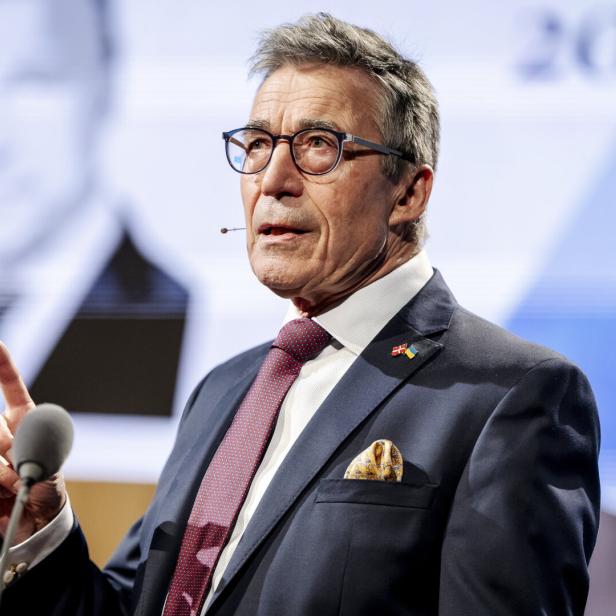Ein Mann mit Brille und Anzug gestikuliert vor einem Mikrofon auf einer Bühne.