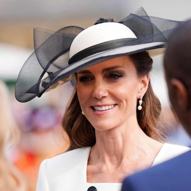 Kate trägt einen eleganten Hut mit schwarzer Schleife und ein weißes Kleid mit schwarzen Knöpfen.