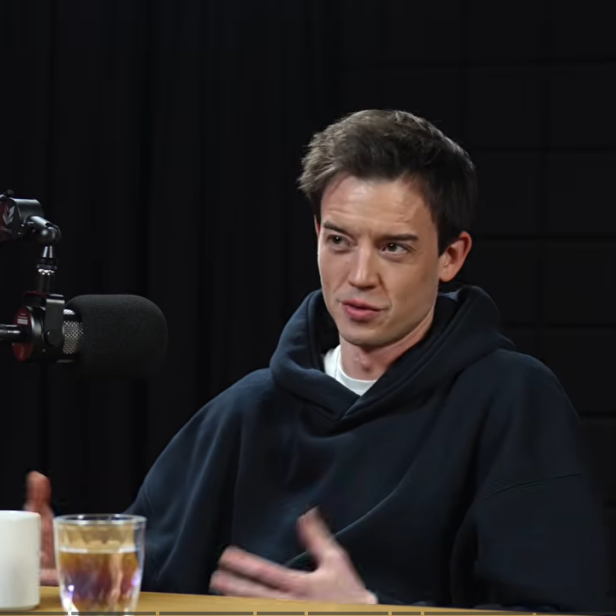 Mann spricht in einem Tonstudio vor einem Mikrofon, auf dem Tisch stehen eine Tasse und ein Glas Wasser.