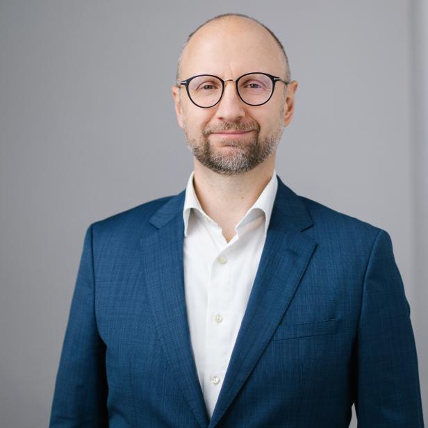 Mann mit Brille und Bart im blauen Anzug vor grauem Hintergrund.