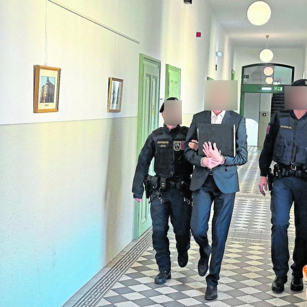 Zwei Justizbeamte führen eine Person in Handschellen durch einen hellen Flur mit grünen Türen und Bildern an der Wand.