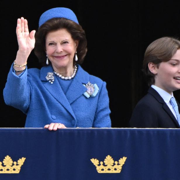 Königin Silvia winkt, begleitet von einem jungen Mann, von einem Balkon.