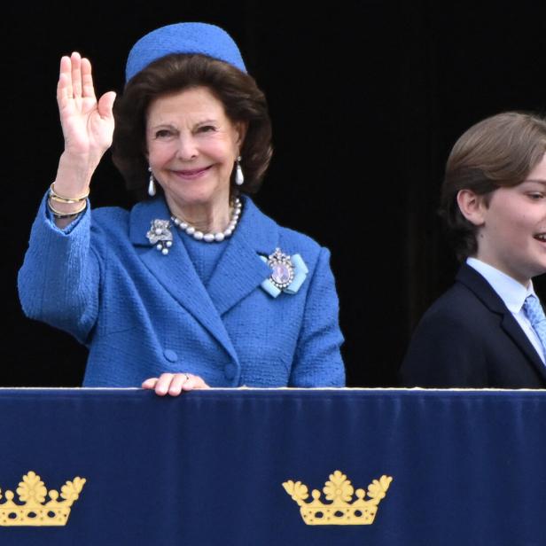 Königin Silvia winkt, begleitet von einem jungen Mann, von einem Balkon.